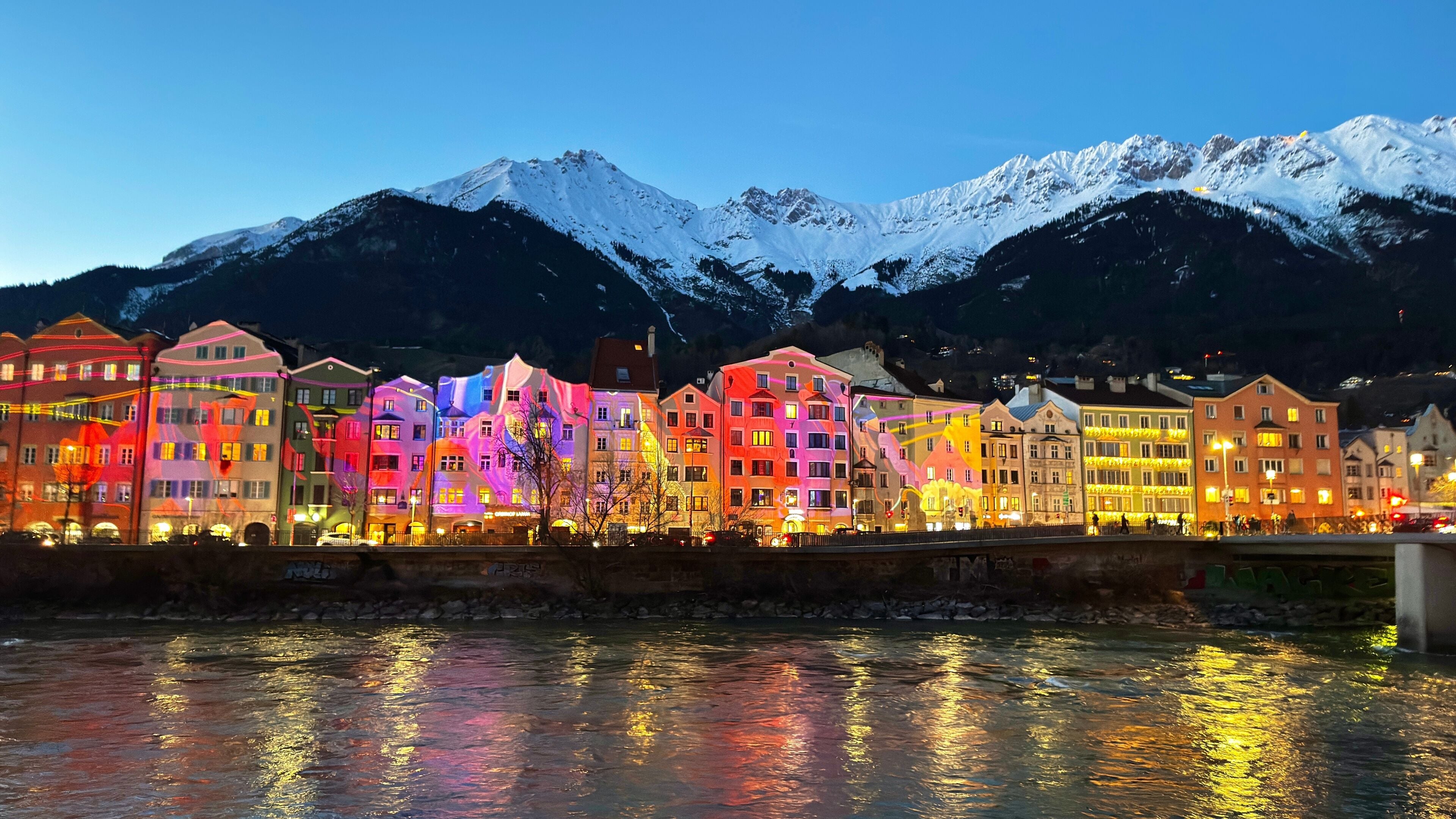 A river reflecting colorful buildings, with snow-capped mountains in the background.