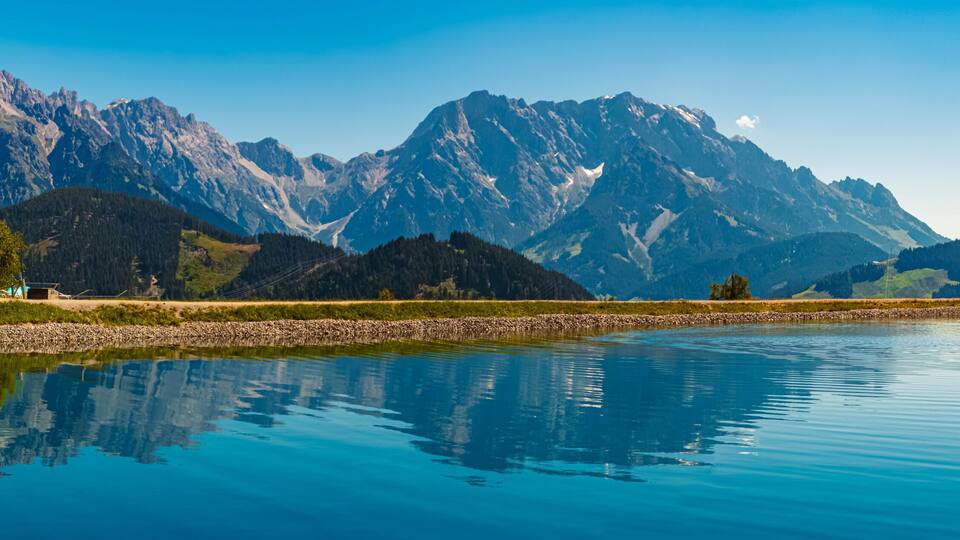A calm lake reflecting mountains, a dock, and a small building.