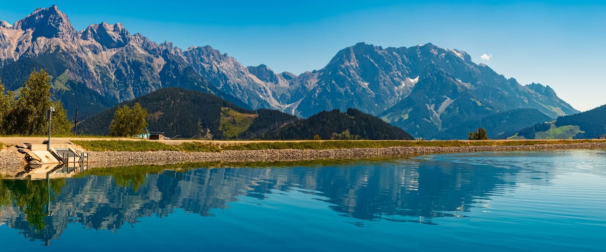 A calm lake reflecting mountains, a dock, and a small building.