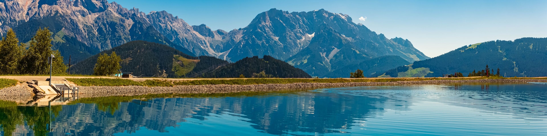 A calm lake reflecting mountains, a dock, and a small building.