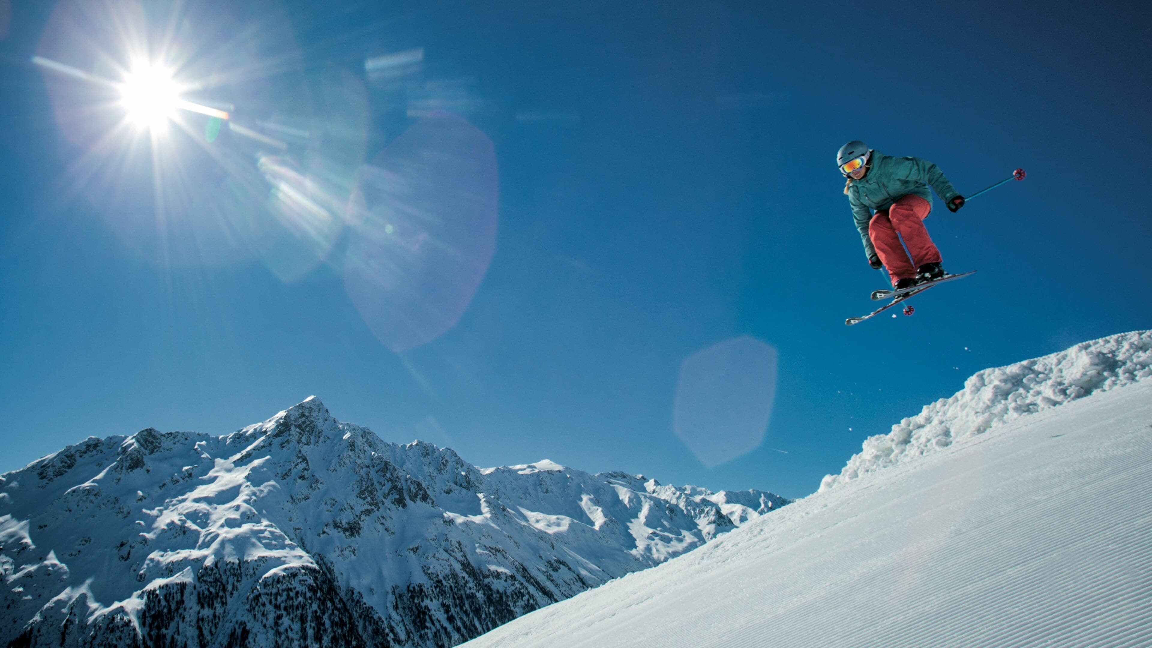 A skier mid-air against a clear blue sky, with snow-covered mountains in the background.