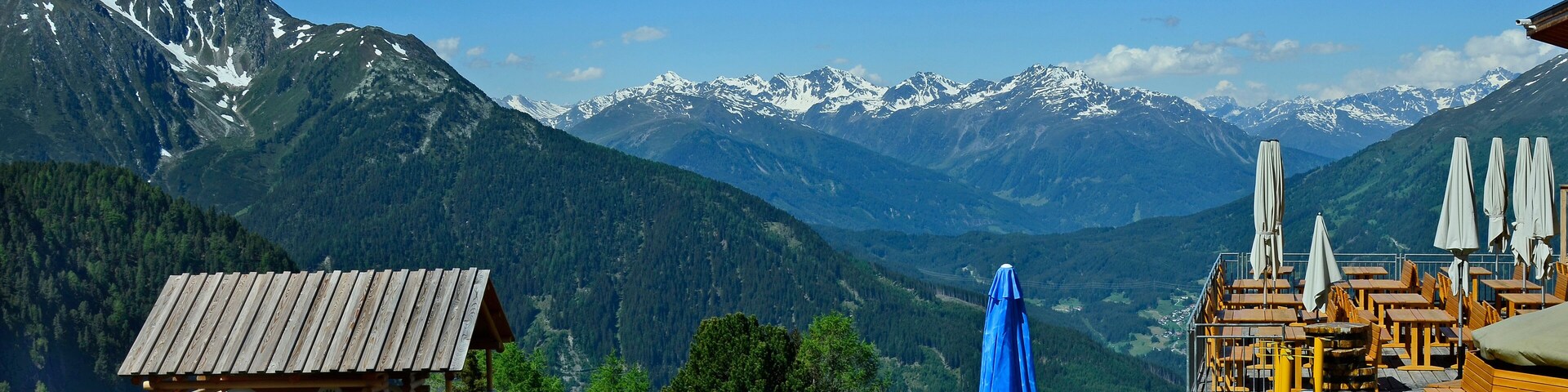 Zona de juegos de madera con arena y juguetes, con vistas a montañas nevadas y un cielo azul despejado.