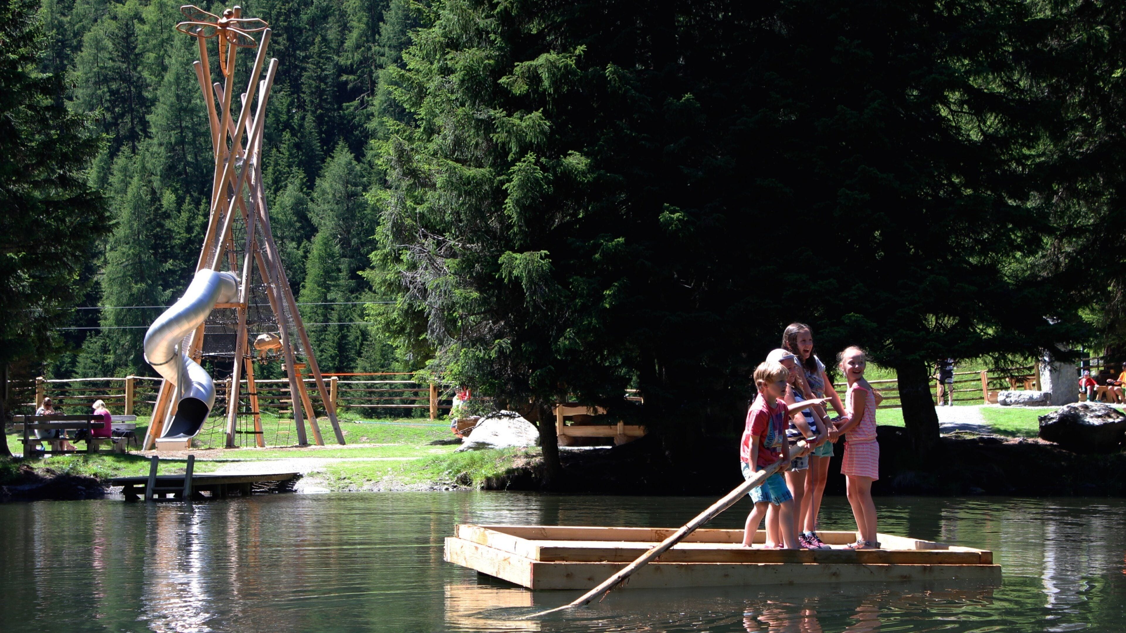 A playground with a slide and swings, a wooden dock over water, and dense forest in the background.