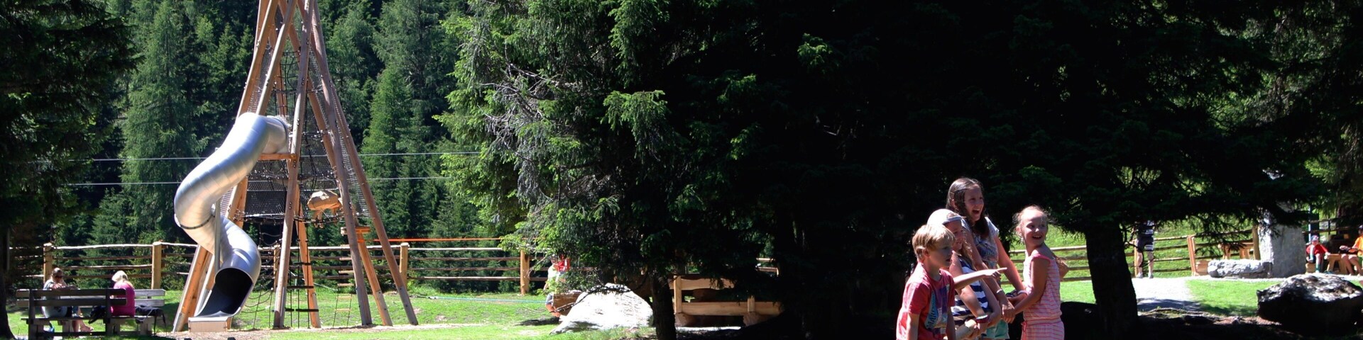 A playground with a slide and swings, a wooden dock over water, and dense forest in the background.