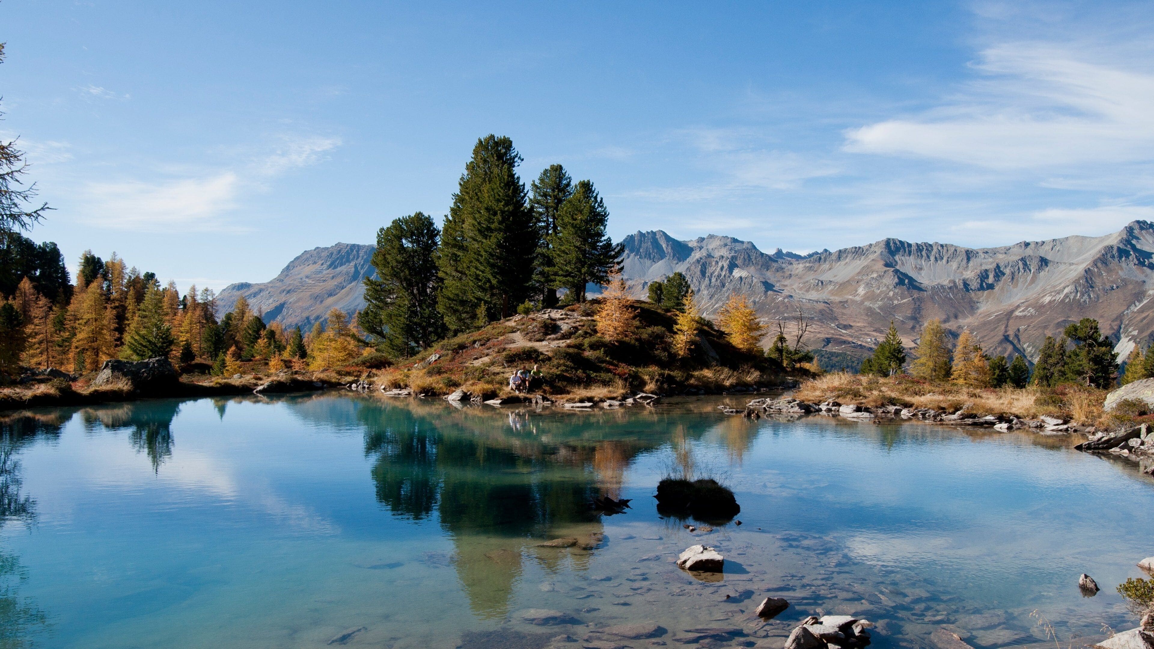 Um lago de montanha límpido, rodeado por árvores de outono e terreno rochoso.