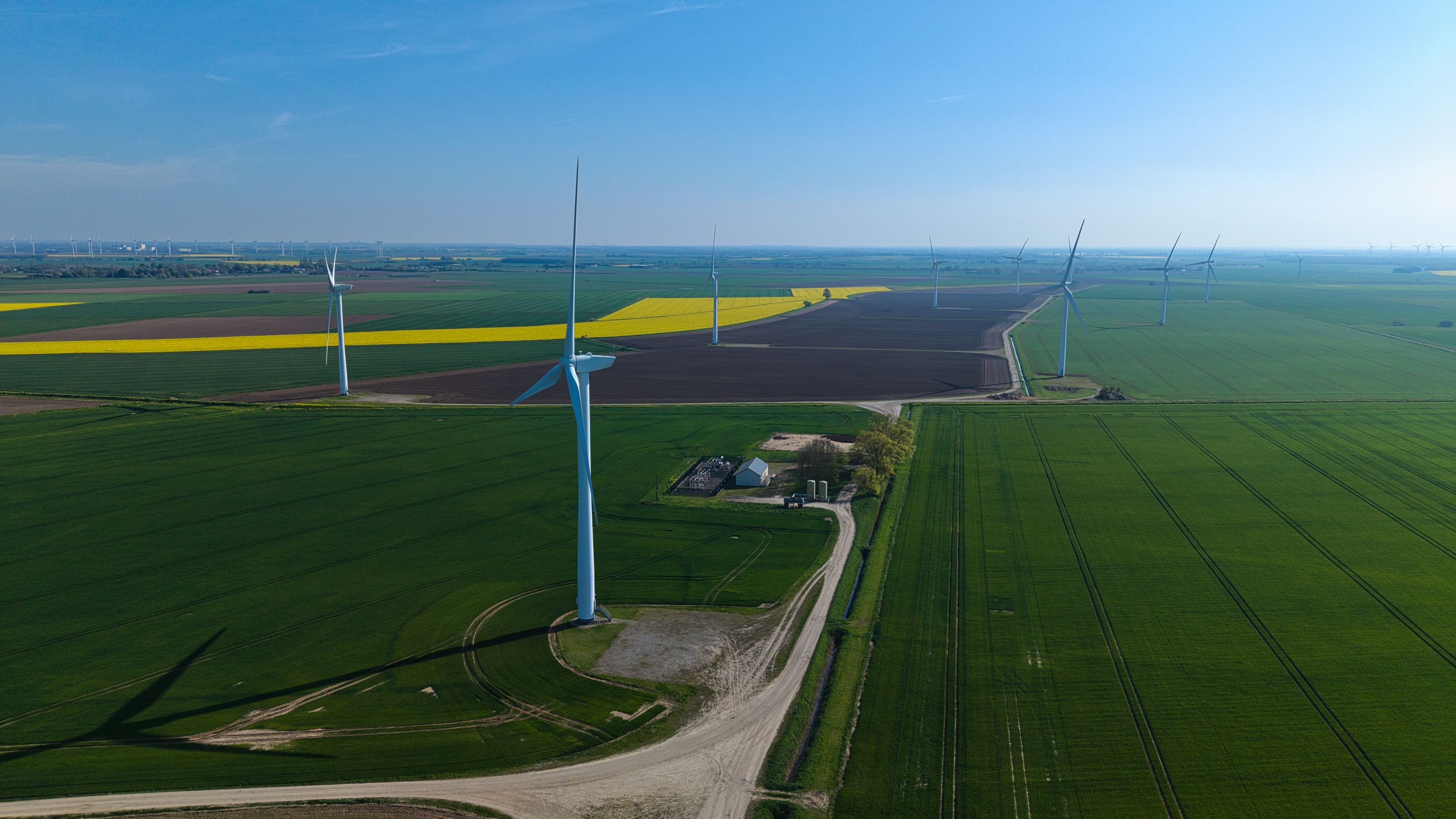 Aerial view of wind turbines standing in vast agricultural fields with vibrant yellow rapeseed and green crops under a clear blue sky in Eastoft, England, United Kingdom.