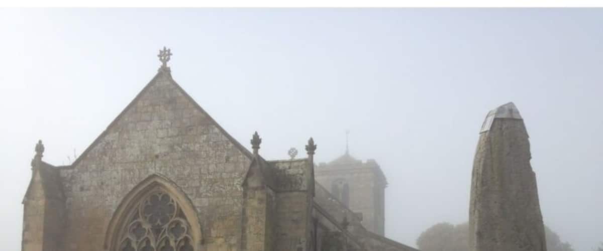 A Gothic church with a pointed roof and large arched windows, surrounded by a foggy cemetery with gravestones.