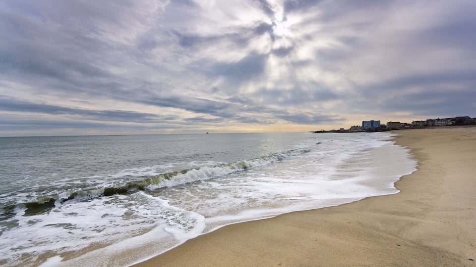 A beach with waves, a cloudy sky, and buildings in the distance.