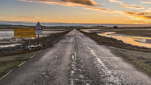 A rural road with warning signs, water on the sides, and a sunset in the distance.