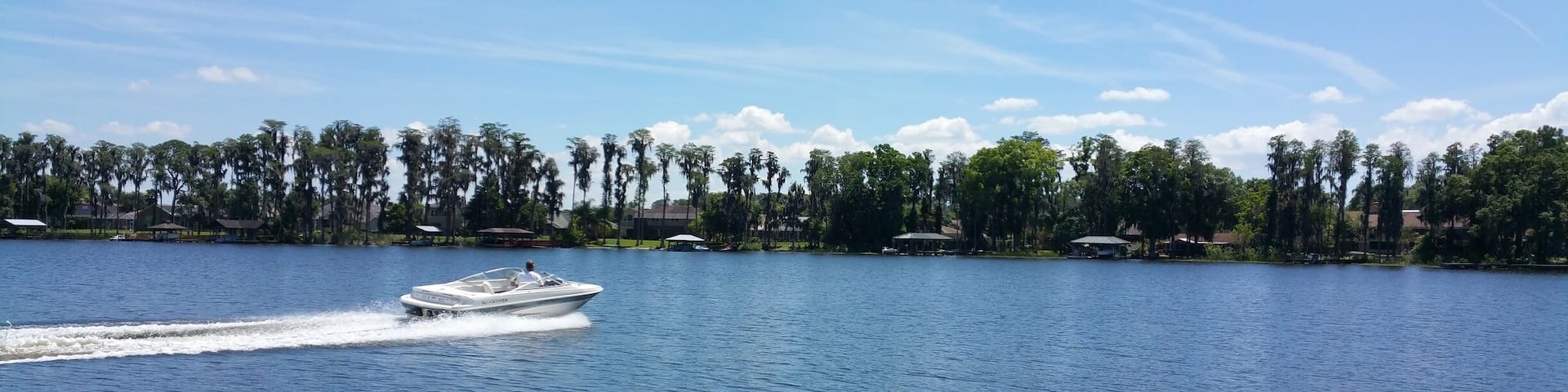 A boat on a calm lake with a clear sky and a line of trees and buildings in the background.