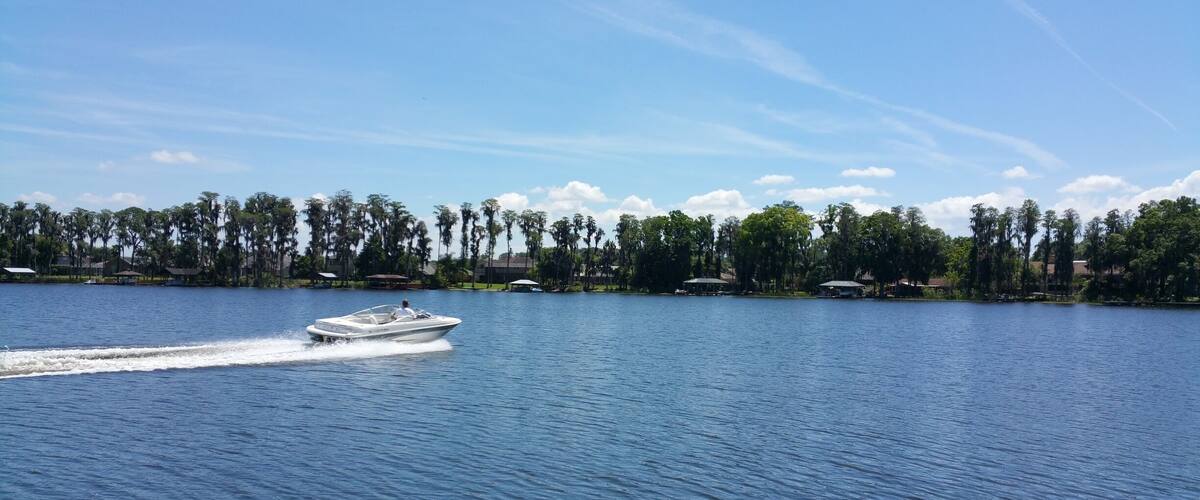 A boat on a calm lake with a clear sky and a line of trees and buildings in the background.