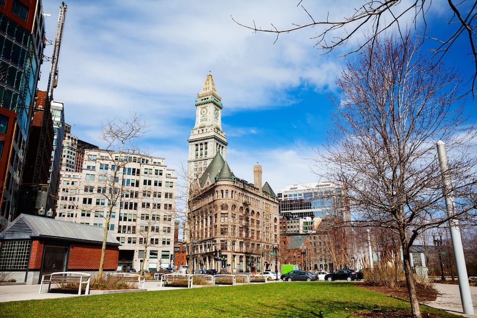 A historic building with a clock tower, surrounded by modern high-rise buildings and a park with a bench.