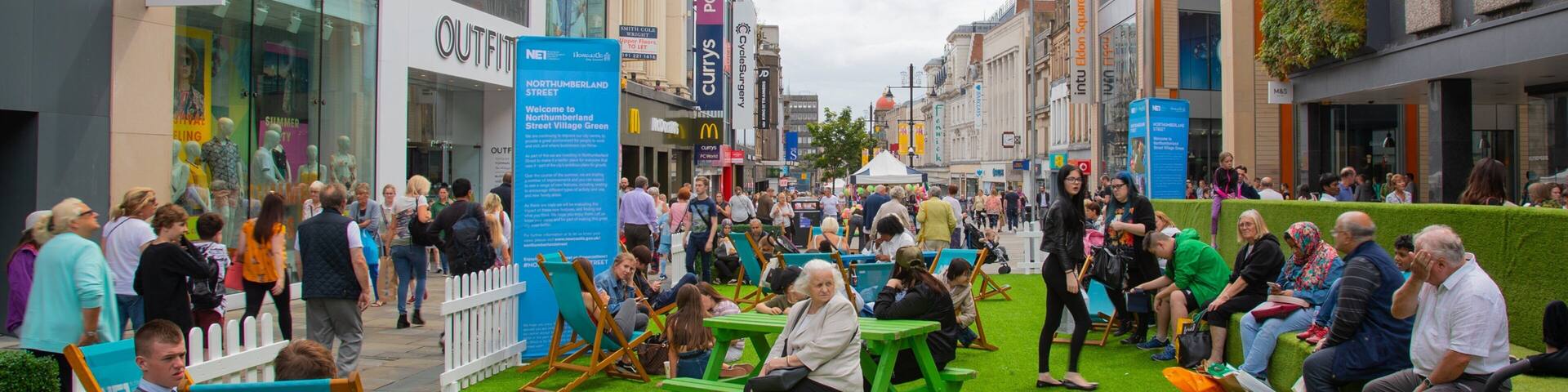 A bustling street with people sitting on green picnic tables, surrounded by shops and buildings.