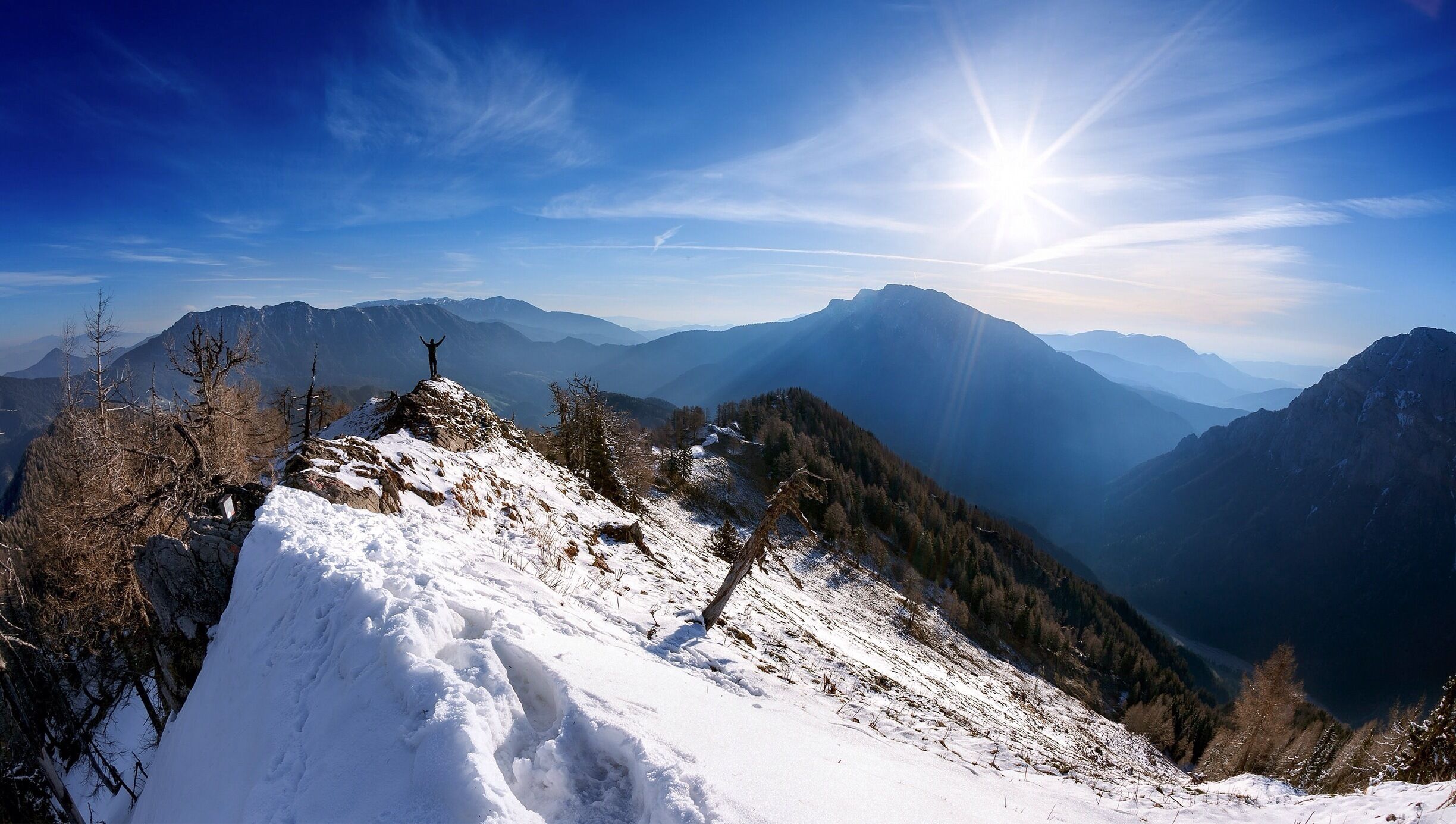 A snowy mountain peak with a person standing on top, surrounded by forested slopes and distant mountain ranges under a clear sky.