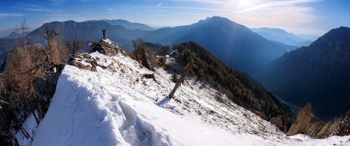 A snowy mountain peak with a person standing on top, surrounded by forested slopes and distant mountain ranges under a clear sky.