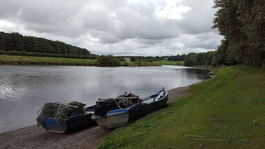 Um rio com barcos e redes de pesca na margem, rodeado por vegetação e um céu nublado.