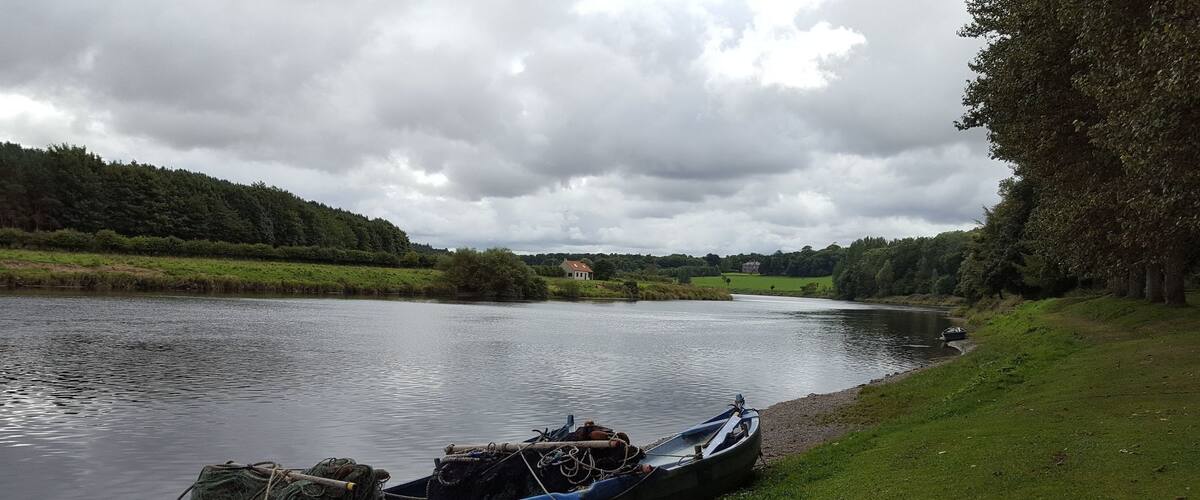 A river with boats and fishing nets on the shore, surrounded by greenery and a cloudy sky.