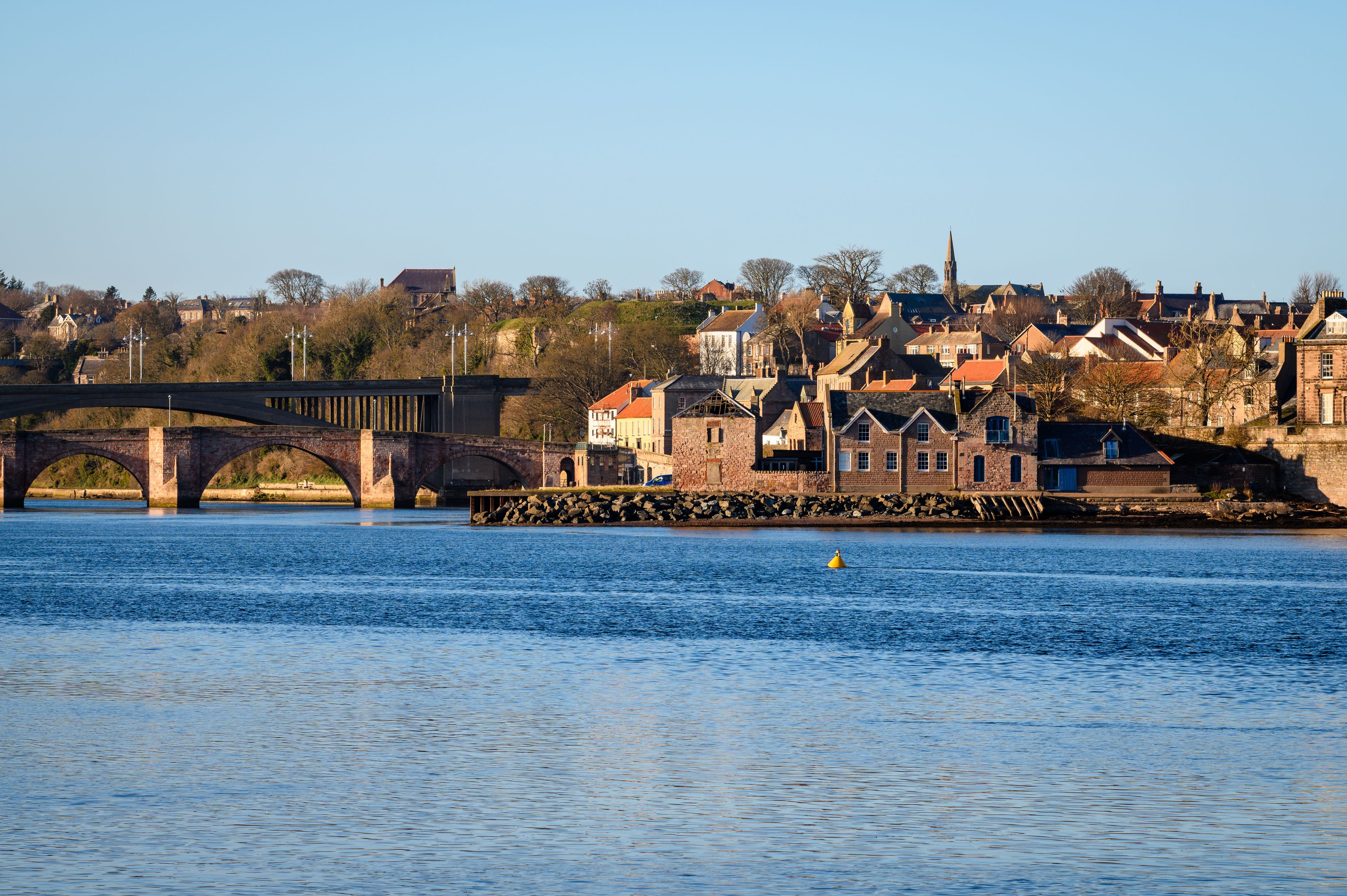 A river with a stone bridge, historic buildings, and a church spire in the background.