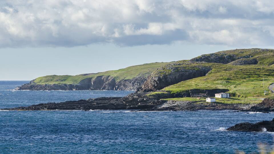 A coastal landscape with rocky cliffs, grassy hills, and a small white building near the shore.