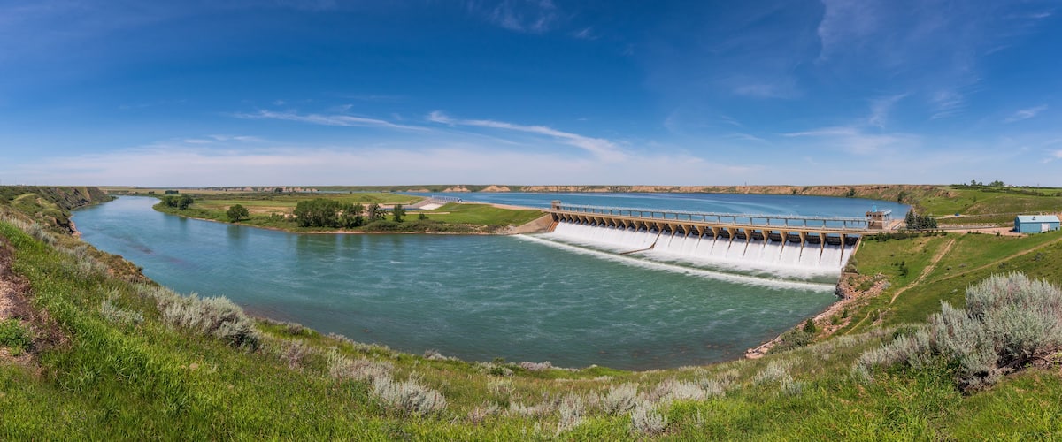 A dam with a spillway releasing water into a river, surrounded by green hills and a clear blue sky.