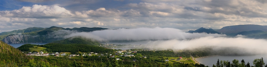 Un pueblo costero rodeado de montañas y bosques densos, con un lago y picos cubiertos de niebla.