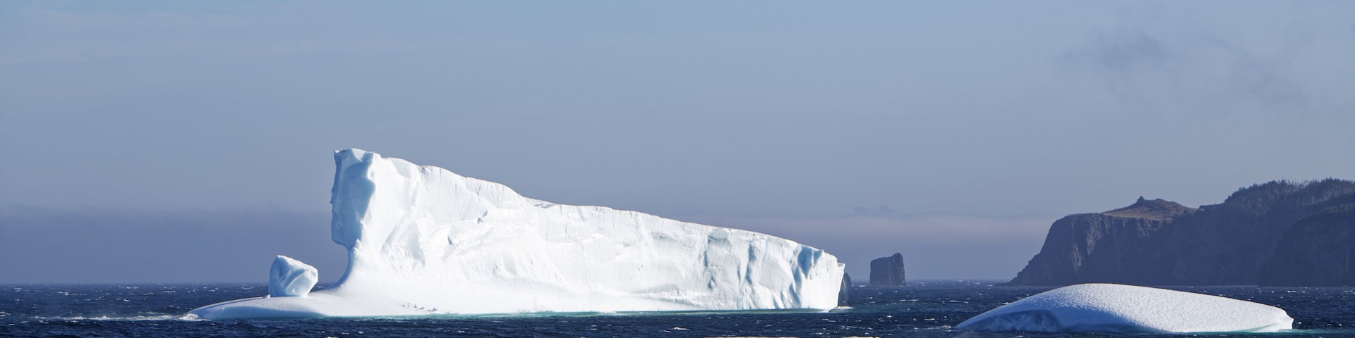 Un iceberg grande en el océano, con icebergs más pequeños cerca y una costa rocosa al fondo.