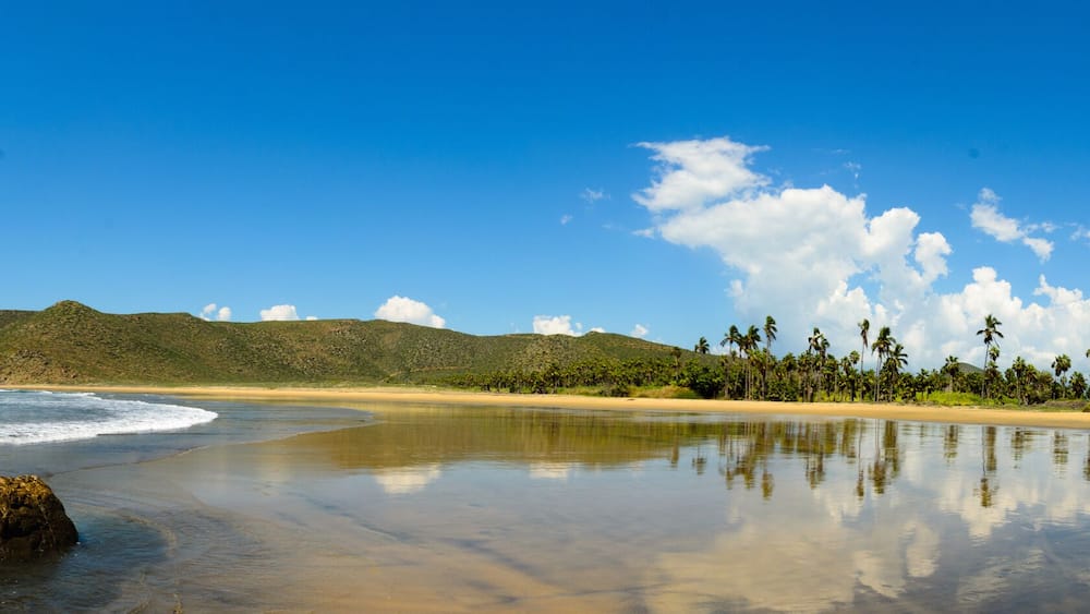 A beach with clear blue water, a sandy shore, a rock in the water, and a mountain in the background.