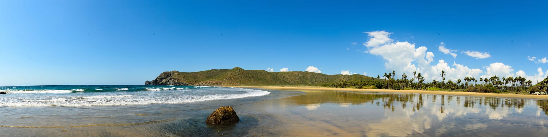 A beach with clear blue water, a sandy shore, a rock in the water, and a mountain in the background.