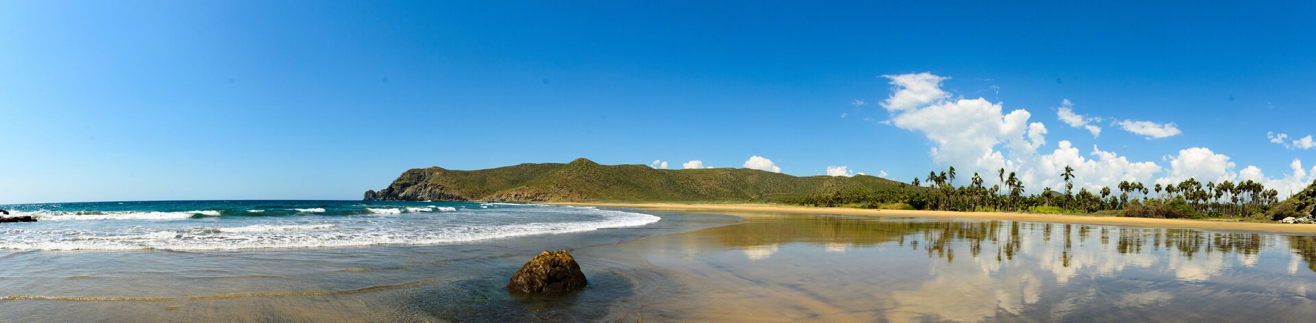 Una playa con agua azul cristalina, orilla arenosa, una roca en el agua y una montaña al fondo.