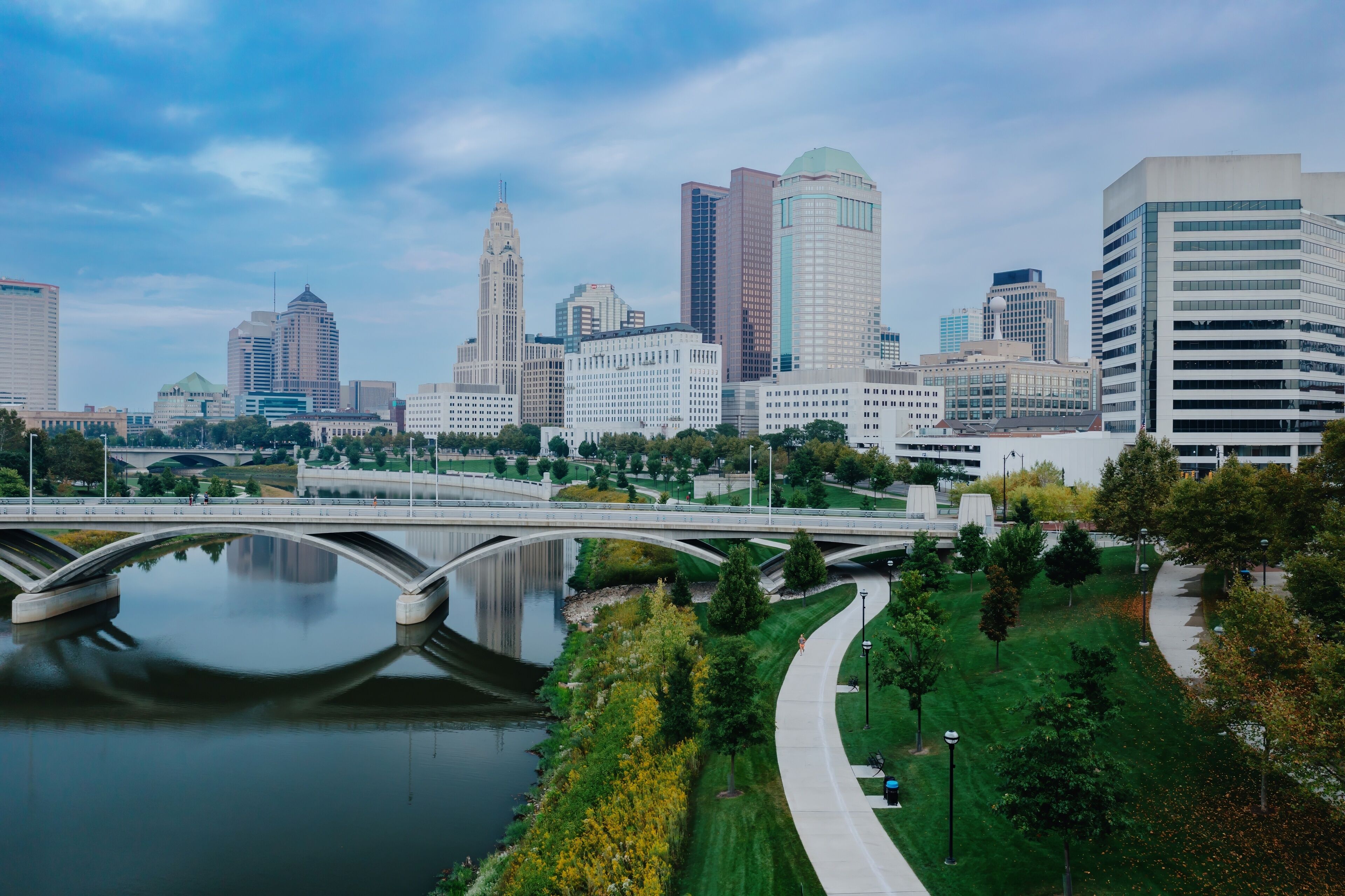 Runner on paved path, city skyline reflected in river, urban park. EAST BANK PARK, COLUMBUS, OHIO, UNITED STATES