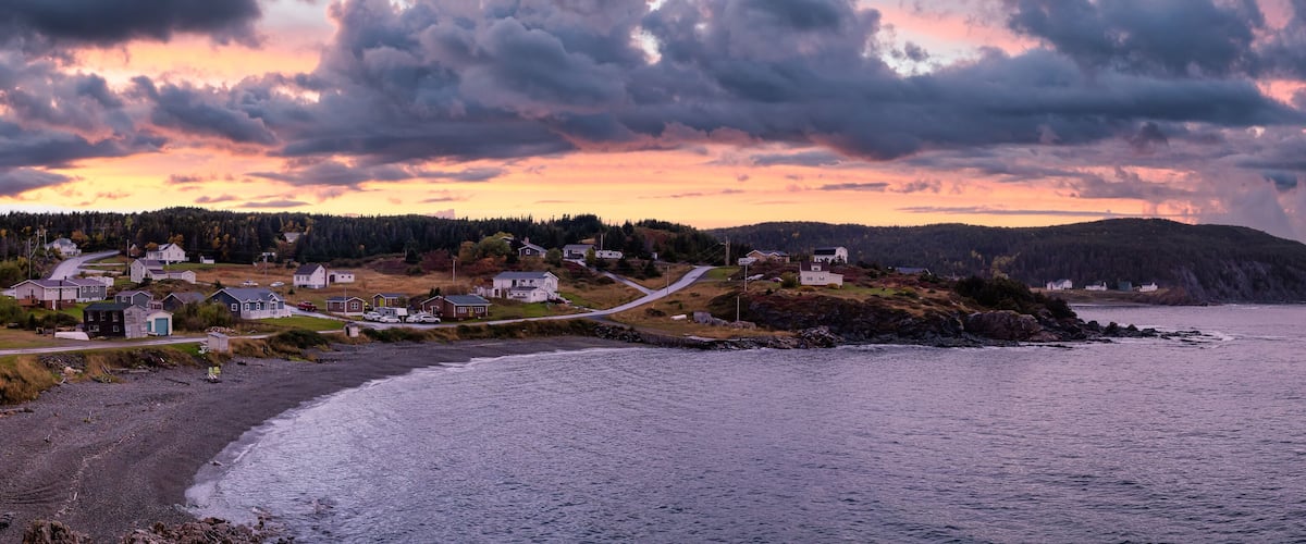 A coastal village with houses along the shoreline, a rocky beach, and a forested hill in the background.