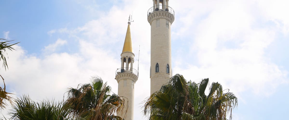 Two tall minarets with golden tips, surrounded by palm trees against a partly cloudy sky.