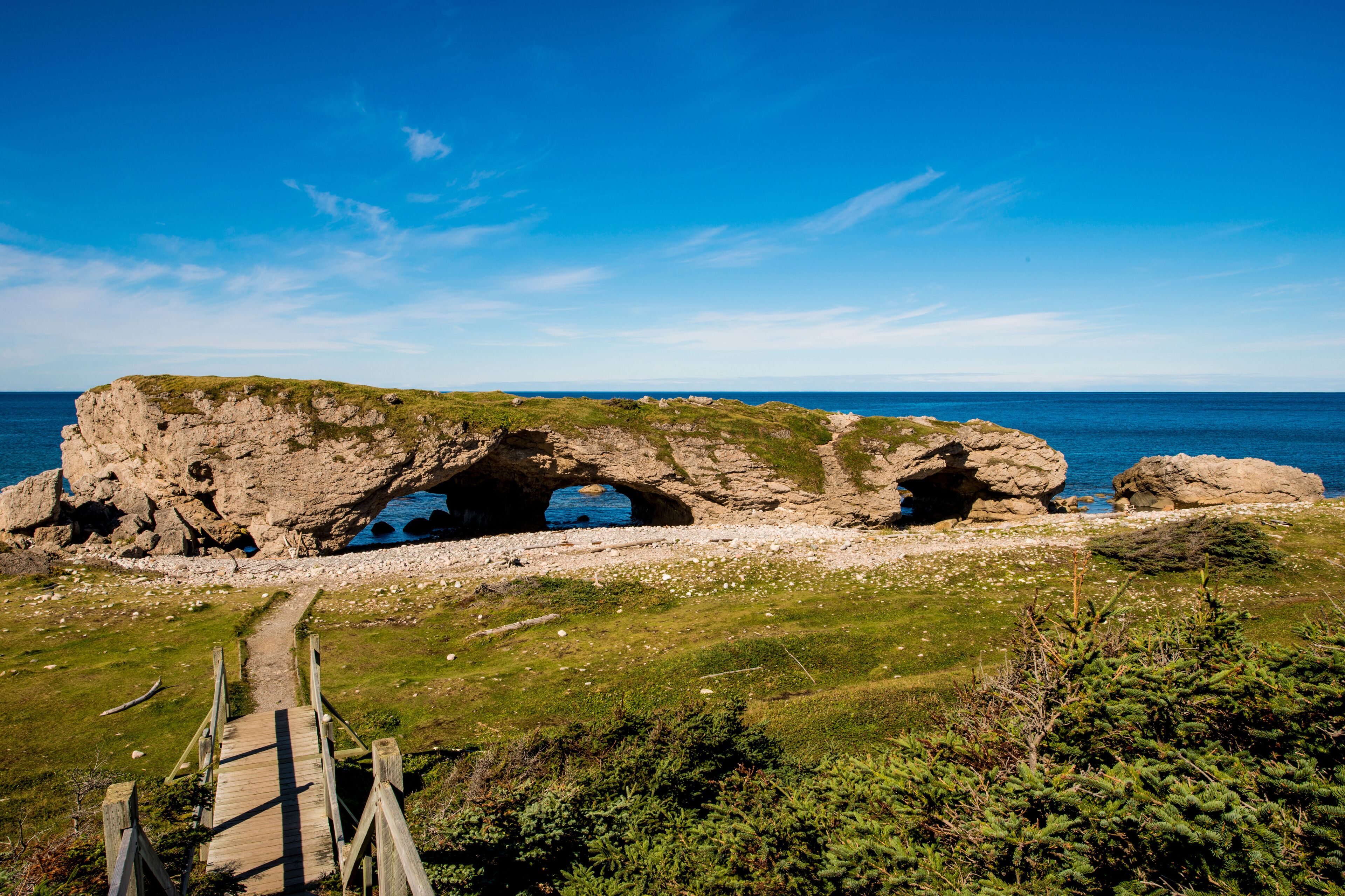 A coastal landscape with a natural rock arch formation, a wooden pathway leading to it, and the sea in the background.