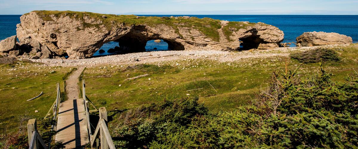 A coastal landscape with a natural rock arch formation, a wooden pathway leading to it, and the sea in the background.
