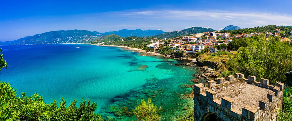 A coastal town with clear turquoise waters, a stone fort, and a backdrop of green hills.