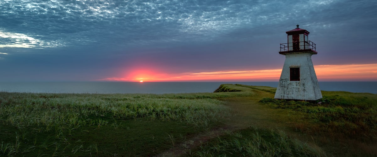 A white lighthouse on a grassy hill with a sunset in the background.