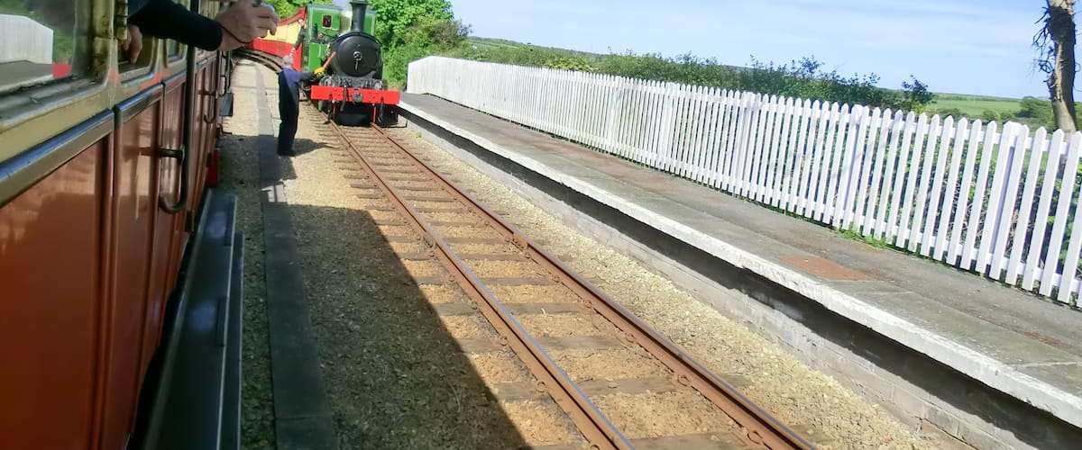 A small train on tracks with a white fence and trees in the background.