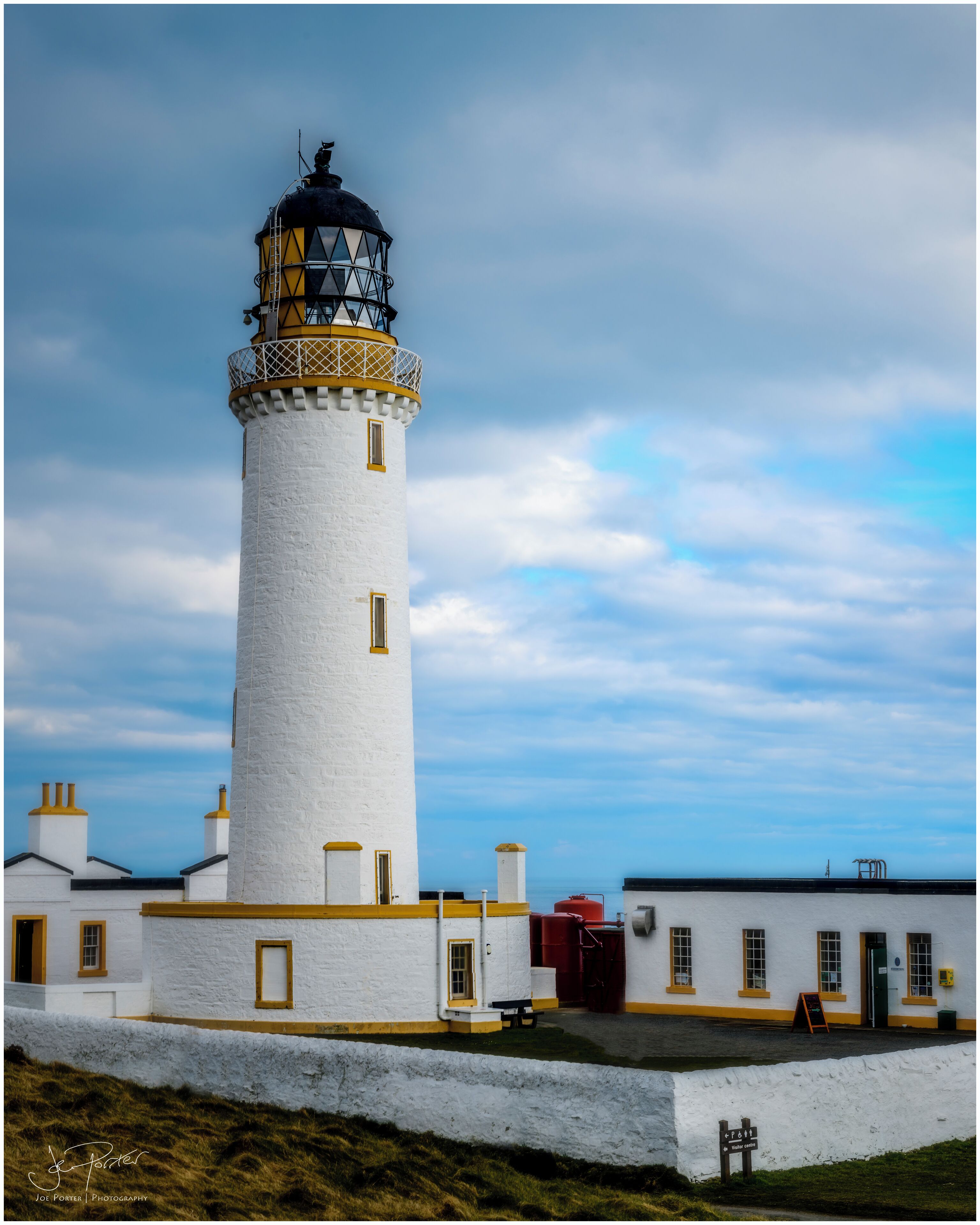 A white lighthouse with a black top and yellow trim, surrounded by white buildings with yellow trim and a grassy foreground.