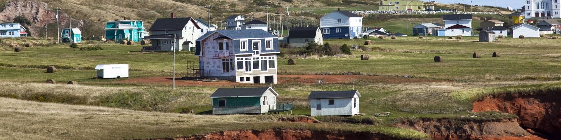 A coastal landscape with houses, a beach, and a cliff with red rock formations.