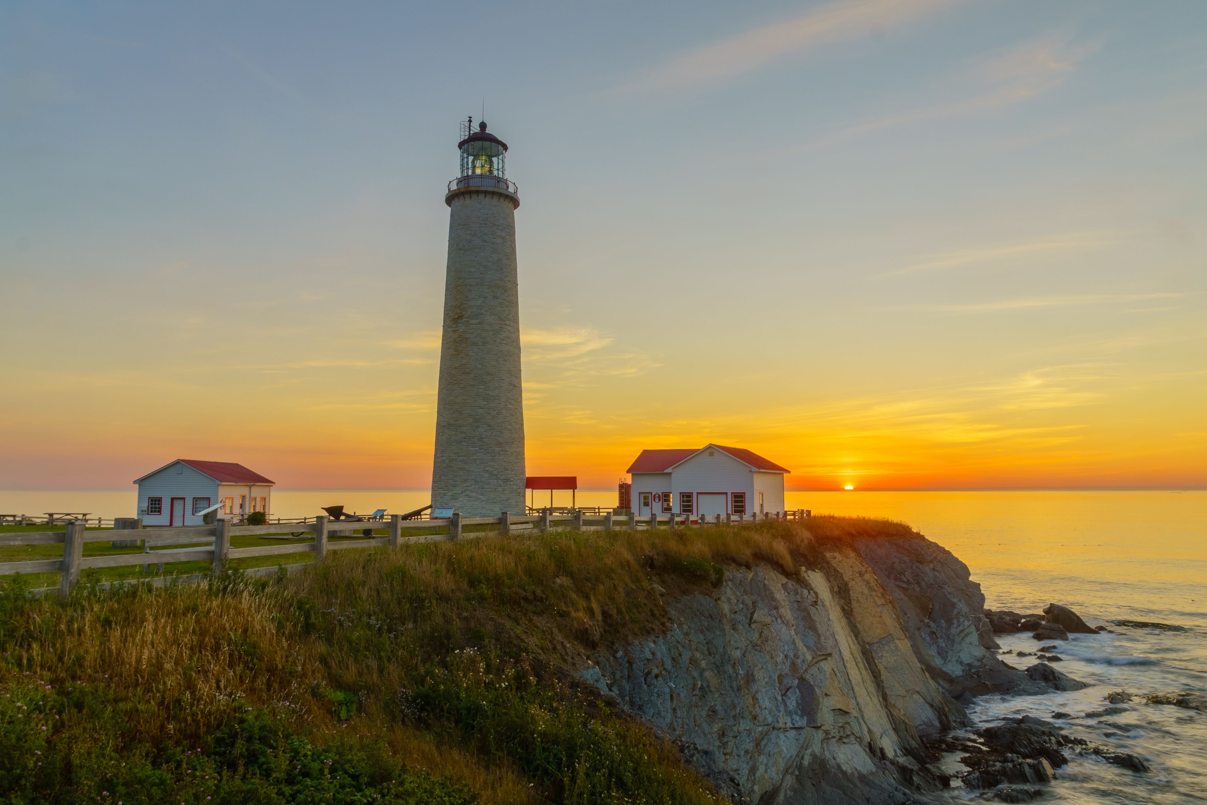 A lighthouse, a wooden structure, and a grassy cliff at sunset.