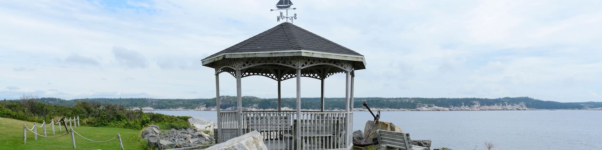 A wooden gazebo with a weather vane, a wooden deck, and a rocky shoreline with a view of the water.