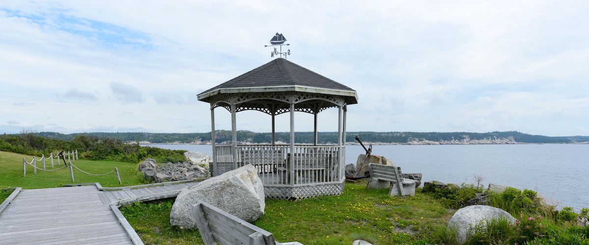 A wooden gazebo with a weather vane, a wooden deck, and a rocky shoreline with a view of the water.