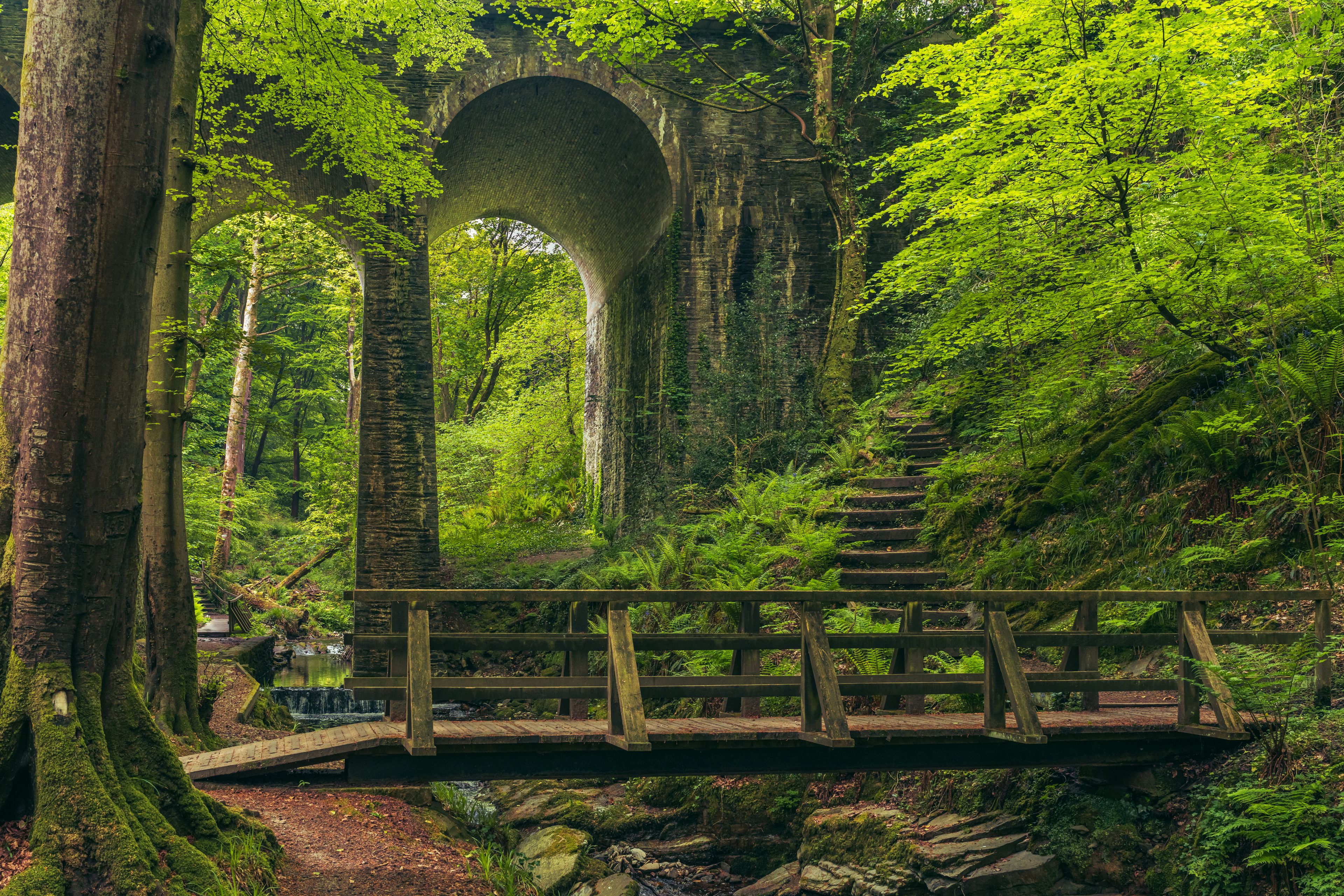 Eine hölzerne Brücke über einem Bach in einem Wald, im Hintergrund eine Brücke mit Steinbogen.