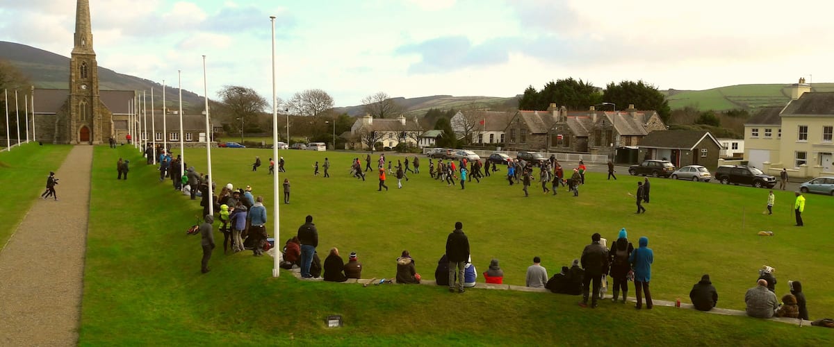 A grassy field with people walking and sitting, a church with a tall steeple, and houses in the background.