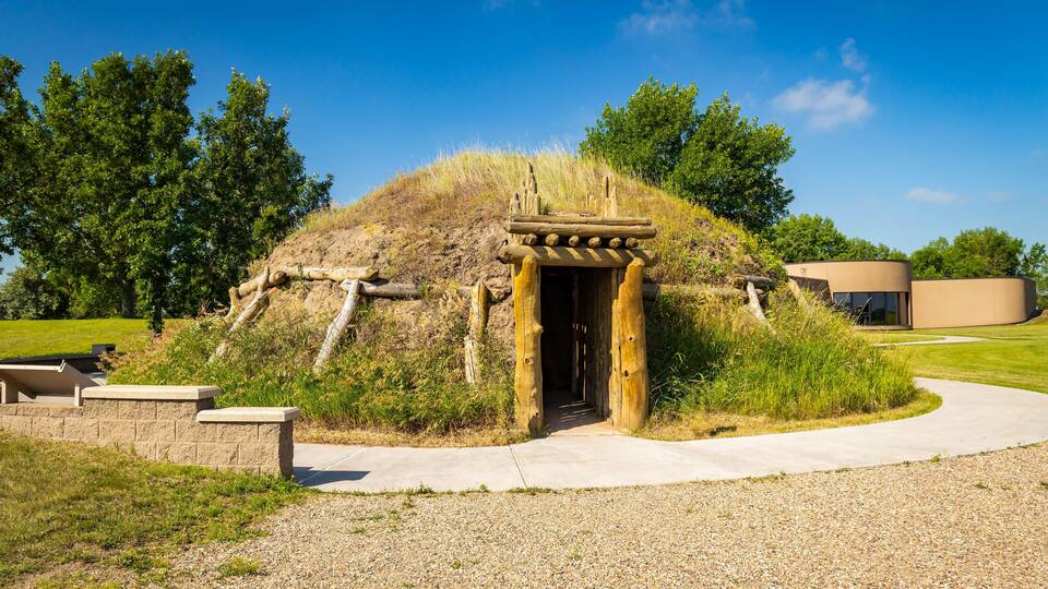 A mound with a wooden entrance, a concrete pathway, and a modern building in the background.