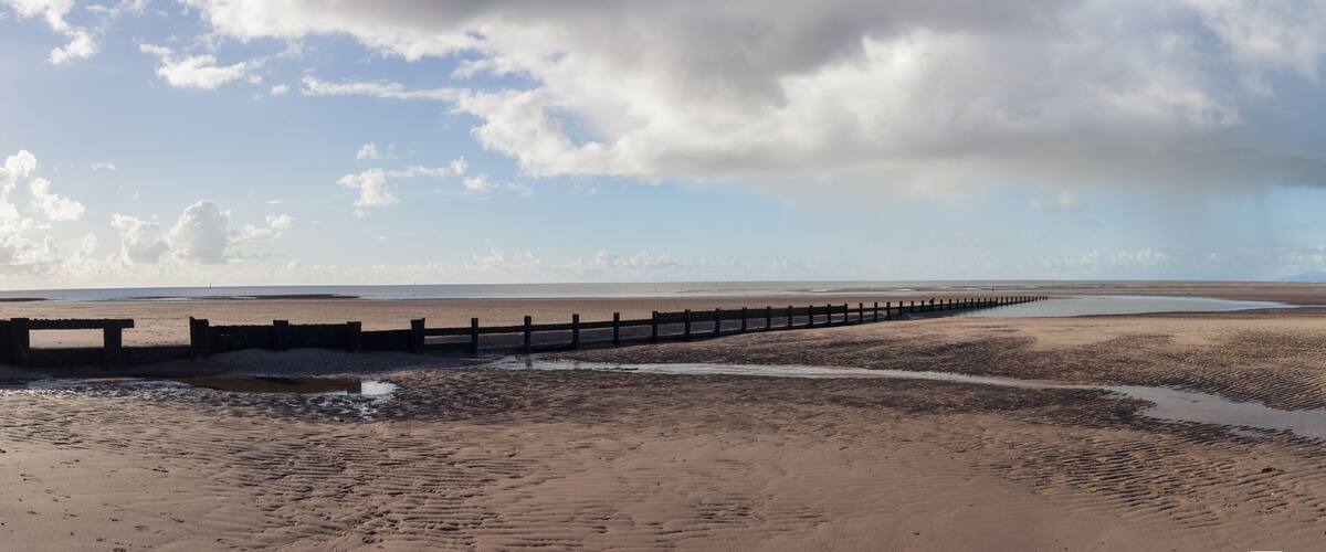 A coastal landscape with a wooden pier extending into a sandy beach, under a partly cloudy sky.