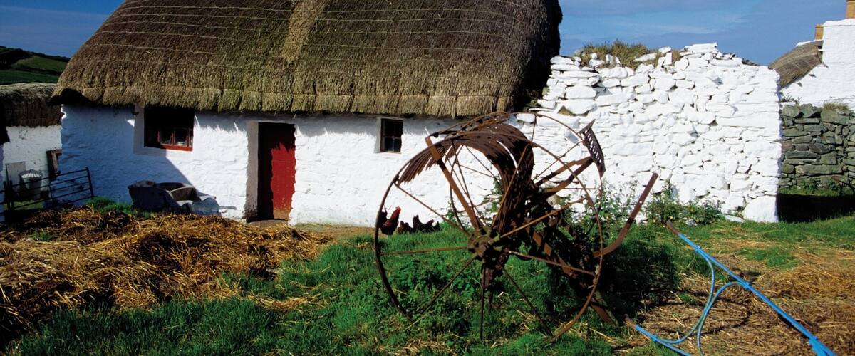 A traditional white house with a thatched roof, a stone wall, and a large metal water wheel.