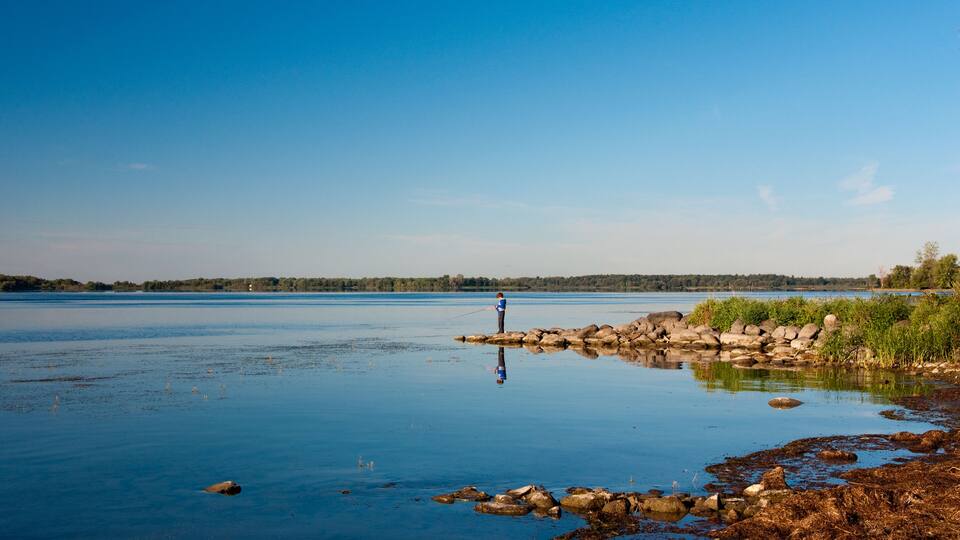 A calm lake with a person fishing on a rocky shore.