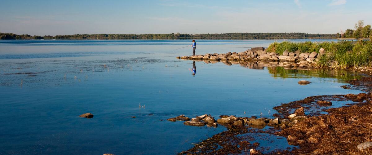 A calm lake with a person fishing on a rocky shore.
