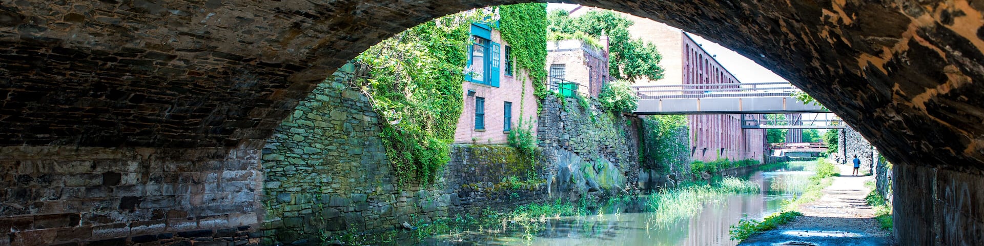 A stone bridge arching over a calm canal, with buildings and greenery in the background.